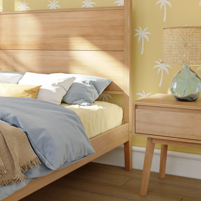 Coastal bedroom close-up of a light wood bed frame and nightstand with woven rattan lamp, soft blue and yellow bedding, and palm-print yellow wallpaper