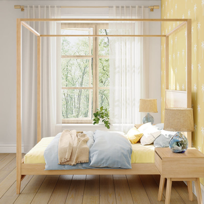 Scandinavian-style bedroom with light wood four-poster canopy bed, pastel blue and yellow bedding, sheer curtains, palm-print yellow accent wall and rattan bedside lamp