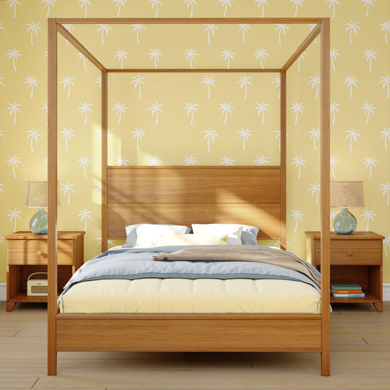 Sunlit coastal bedroom with a natural wood four-poster canopy bed, yellow palm-print wallpaper, matching nightstands and rattan table lamps