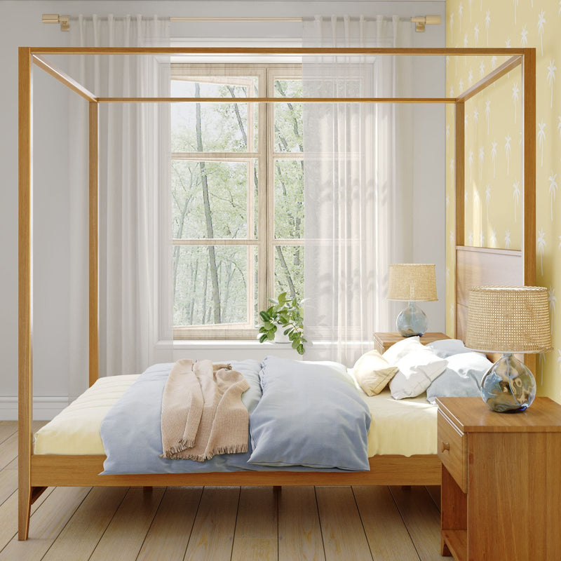 Cozy bedroom with light-wood four-poster canopy bed, pastel blue and beige bedding, sheer curtains over a sunlit window, bedside table with ceramic lamp, potted plant, and yellow palm-print wallpaper