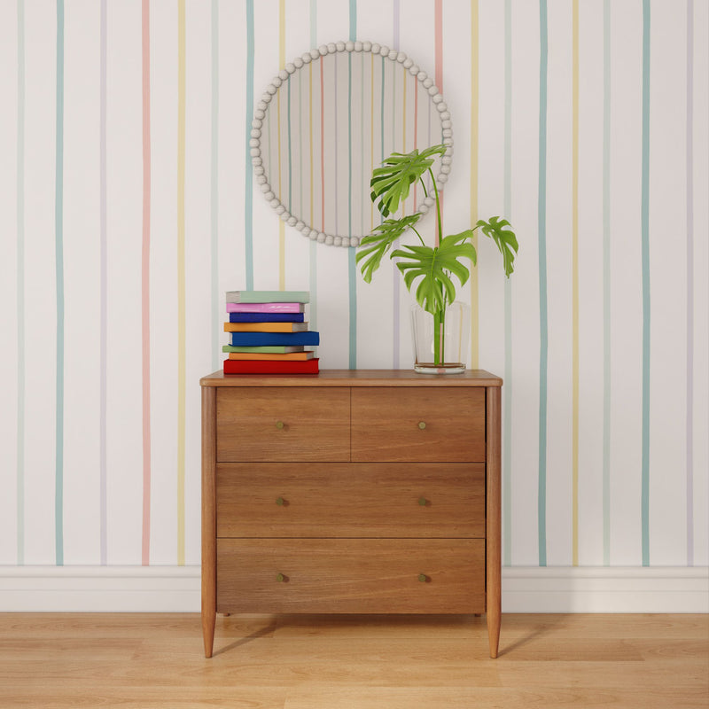 Mid-century modern wooden dresser styled with stacked colorful books, glass vase with monstera leaves, round beaded mirror, and pastel striped wallpaper on a hardwood floor