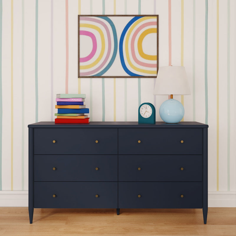 Navy blue six-drawer dresser against pastel striped wallpaper with abstract rainbow wall art, blue table lamp, teal clock and stack of colorful books on wooden floor