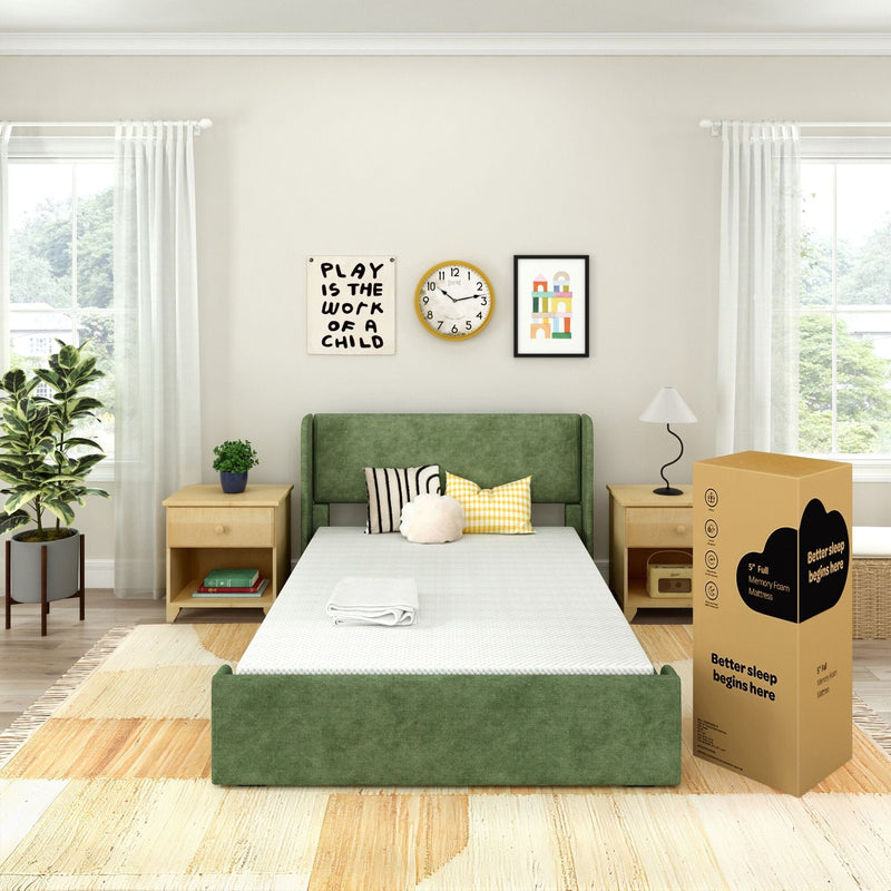 Green upholstered bed with boxed memory foam mattress in a bright modern bedroom with wooden nightstands, lamp, houseplant, decorative pillows and children's wall art