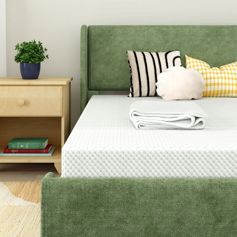 Modern bedroom with green upholstered bed and headboard, white quilted mattress and folded blanket, striped and yellow gingham decorative pillows, wooden nightstand with potted plant and books.
