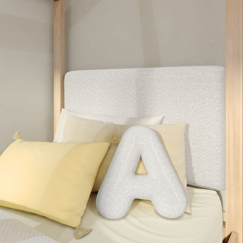 Plush white letter A pillow on cream bedding with soft yellow decorative pillows, textured off-white headboard and light wood canopy frame in a neutral modern bedroom