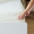 Person adjusting a white fitted sheet on a mattress in a bedroom setting.