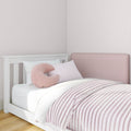 Minimalist nursery toddler bed with pink-and-white striped bedding, plush crescent pillow, white wood frame and pink upholstered headboard