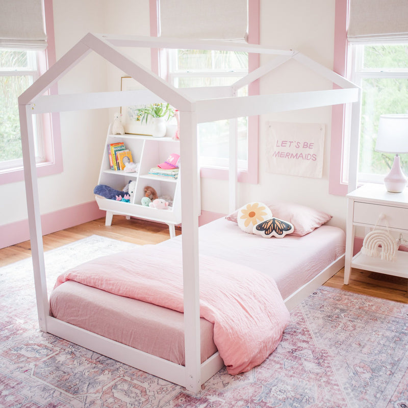 Pastel pink girls' bedroom with white house-frame twin bed, pink bedding, butterfly pillow, toy shelf and "Let's Be Mermaids" wall art