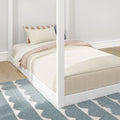 White four-poster bed with beige duvet, striped tan-and-white pillows, light wood floor and blue patterned rug in minimalist bedroom