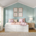 Pastel bedroom with mint-green accent wall and four framed botanical prints, white storage bed with pink floral bedding and pillows, wooden nightstand with table lamp and sheer curtains