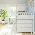 Bright coastal nursery with white crib and dresser, blue wave wallpaper, framed ocean prints, potted plant and sunlit window