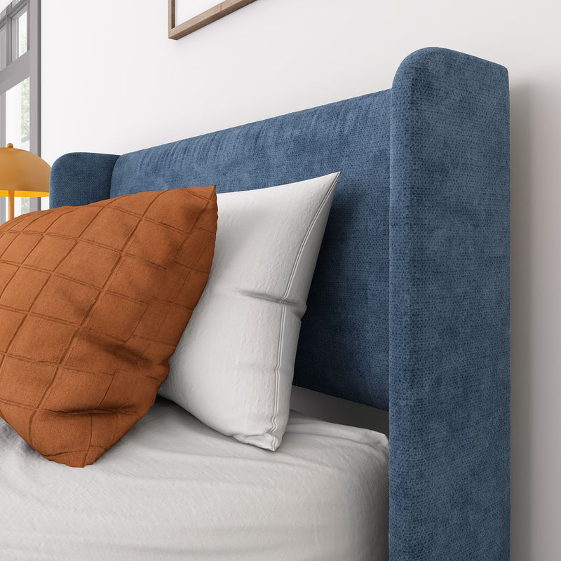 Close-up of blue upholstered headboard with textured fabric, white pillow and rust-orange throw pillow in a modern bedroom interior with lamp
