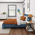 Modern children's bedroom decor with blue upholstered bed, orange quilt and pillows, bedside table with lamp and clock, soccer ball, bookshelf and framed wall art