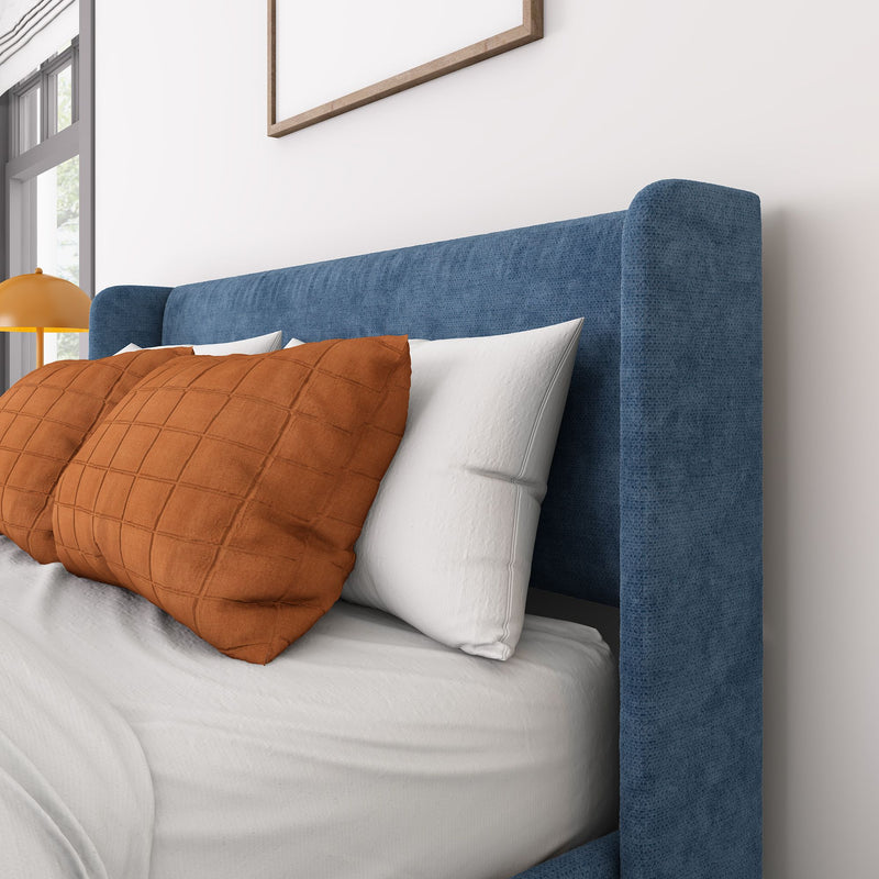 Close-up of a contemporary bedroom with a blue upholstered headboard, orange quilted pillows, white bedding, bedside lamp, window light and minimal wall art