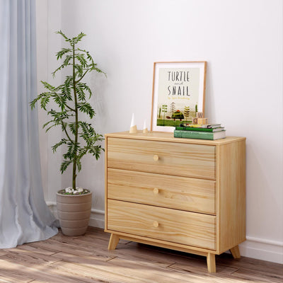 Minimalist light wood three-drawer dresser with mid-century legs styled in a Scandinavian nursery setting — potted green plant, framed 