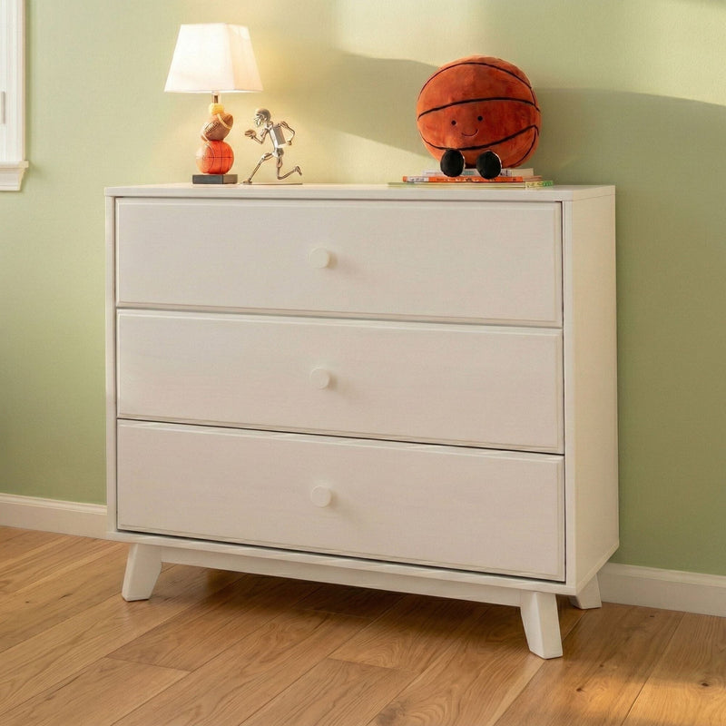 White mid-century modern three-drawer dresser chest of drawers for nursery or bedroom, styled with a lamp and plush basketball on a hardwood floor against a pale green wall