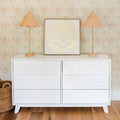 White six-drawer dresser with rattan table lamps, framed gold butterfly art, woven basket on wood floor and floral wallpaper backdrop