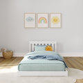 Nursery wall art: three pastel framed cloud, rainbow and sun prints above a white toddler bed with striped bedding, plush sun pillow and wicker toy basket