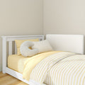 White wooden crib with yellow striped bedding, soft C-shaped nursing pillow and white headboard against a neutral beige nursery wall