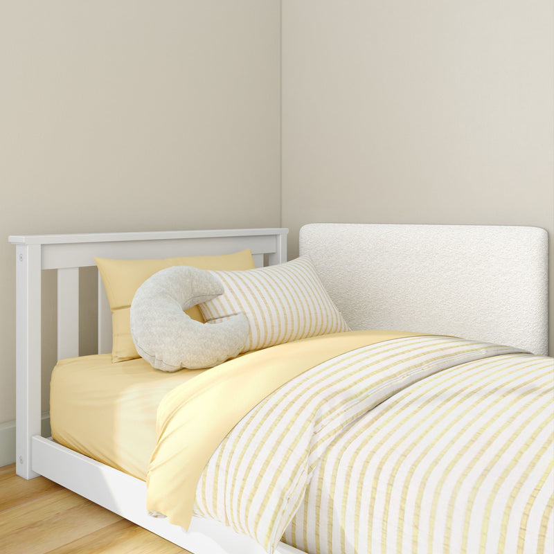 White wooden crib with yellow striped bedding, soft C-shaped nursing pillow and white headboard against a neutral beige nursery wall
