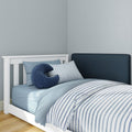 White wooden slatted toddler bed with blue striped bedding, light blue sheets and navy U-shaped pillow against a navy upholstered headboard and pale blue nursery wall, modern children’s bedroom and nursery decor