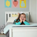 Smiling young girl with white bow lying on a white bed in a modern children's bedroom with pastel bedding and colorful fruit wall art
