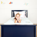 Smiling girl lying on bed in navy blue bed frame with pastel bedding in bright kids bedroom, geometric wall decals and a picture book nearby