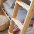 Close-up of natural pine wooden bunk bed ladder in a cozy bedroom with grey linen bedding, woven pouf and pink throw