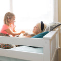 Two smiling young girls playing on a white daybed by a sunny window with a cozy knit blanket and floral dresses
