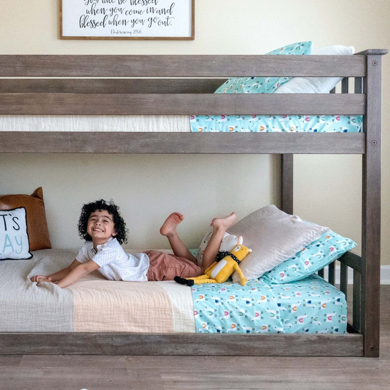 Smiling young boy lying on the lower bunk of a gray wooden kids bunk bed in a bright children's bedroom with teal animal-print bedding, plush toy and farmhouse-style wall art