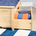 Under-bed storage drawer in natural pine bed containing a rolled blue-and-orange blanket and gray pillow on a blue-and-white striped rug — space-saving bedroom storage solution
