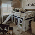 White wooden kids loft bed with slide and ladder, playhouse curtain underneath, neutral boho children's bedroom decor with plaid pillows and woven pendant light by window