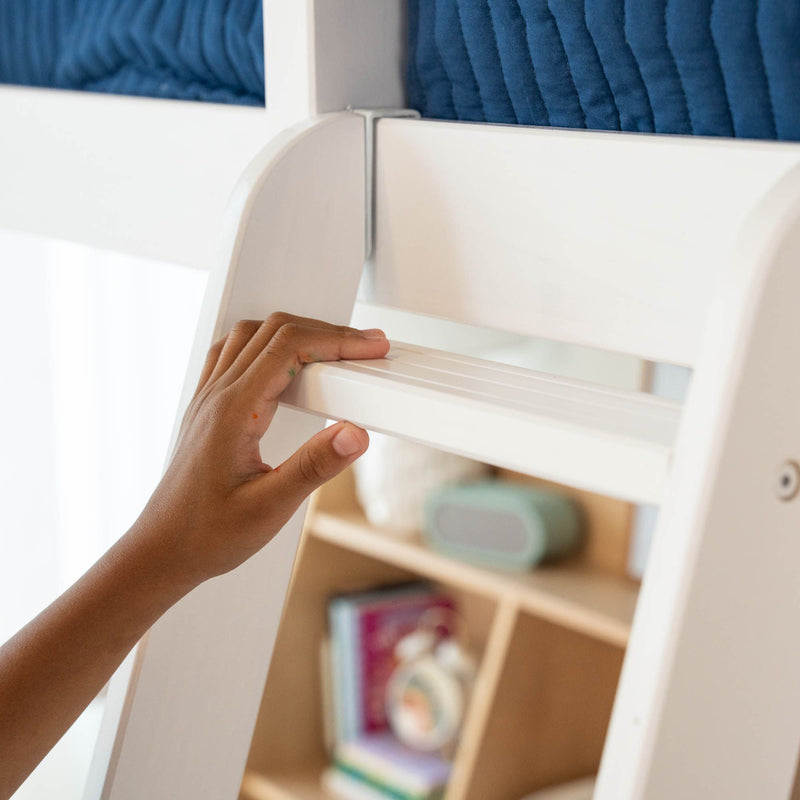 Close-up of a child's hands gripping a white bunk bed ladder in a children's bedroom with blue quilted bedding and a blurred wooden bookshelf
