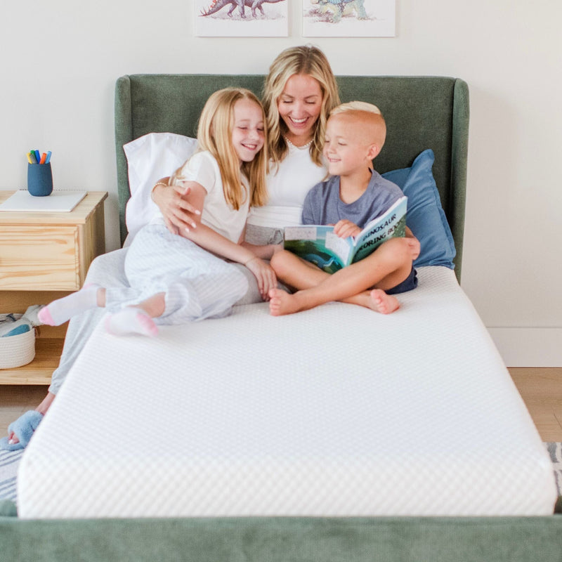 Mother reading a bedtime story to two smiling children on a white mattress in a bright kids' bedroom with a green upholstered headboard, wooden nightstand and dinosaur wall art