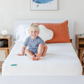 Smiling toddler sitting on a white children's mattress in a bright modern nursery with a rust-colored pillow and wooden nightstands