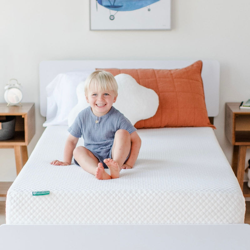 Smiling toddler sitting on a white children's mattress in a bright modern nursery with a rust-colored pillow and wooden nightstands