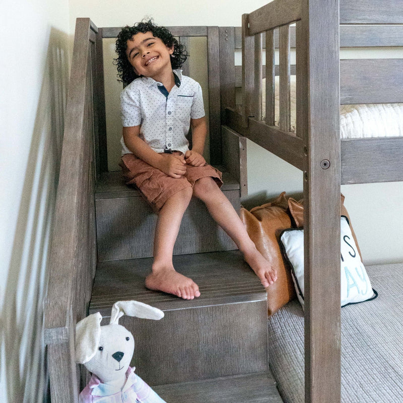 Smiling child sitting barefoot on wooden bunk bed stairs in cozy family bedroom, wearing a white dotted shirt and brown shorts
