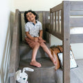 Smiling young boy with curly hair sitting on wooden bunk bed stairs in a bright children's bedroom wearing a white polka-dot shirt and brown shorts, stuffed animal and pillows nearby
