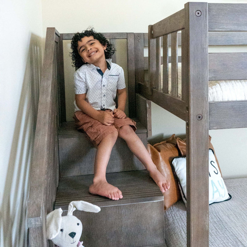 Smiling curly-haired child sitting on wooden bunk bed stairs in a modern kids' bedroom with a plush toy and decorative pillows
