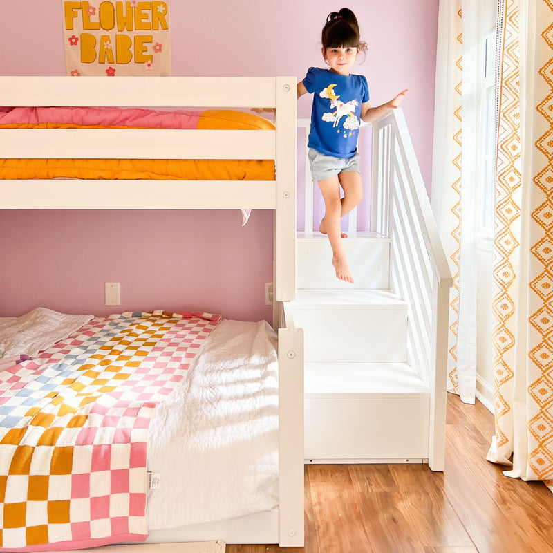 Kids bunk bed with white wooden stairs and little girl in a unicorn t-shirt in a pink children’s bedroom, colorful checkered quilt, orange patterned curtains and "Flower Babe" wall art