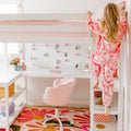 Pink kids' bedroom with white loft bed and ladder, built-in desk, floral rug and photo string lights, child in pink tie-dye pajamas climbing the bed