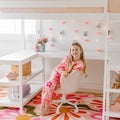 Young girl in pink tie-dye loungewear sitting at a white desk under a loft bed in a pastel kids' bedroom with heart string lights, floral rug, and playful pink decor