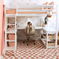 Kids loft bed with built-in desk, white frame and peach bedding, sun-pattern wallpaper, child studying at desk, pink checkered rug, cactus and disco ball decor