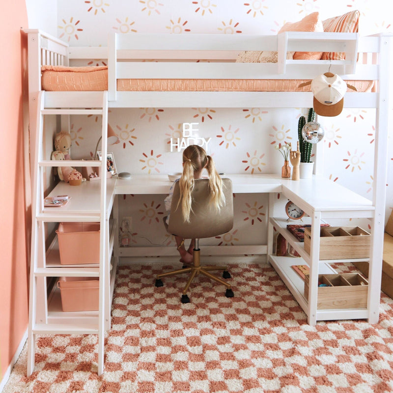Kids loft bed with built-in desk, white frame and peach bedding, sun-pattern wallpaper, child studying at desk, pink checkered rug, cactus and disco ball decor