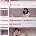 Kids bunk beds: two smiling girls lounging on white wooden bunk beds with floral bedding, pink wallpaper and cozy children's room decor