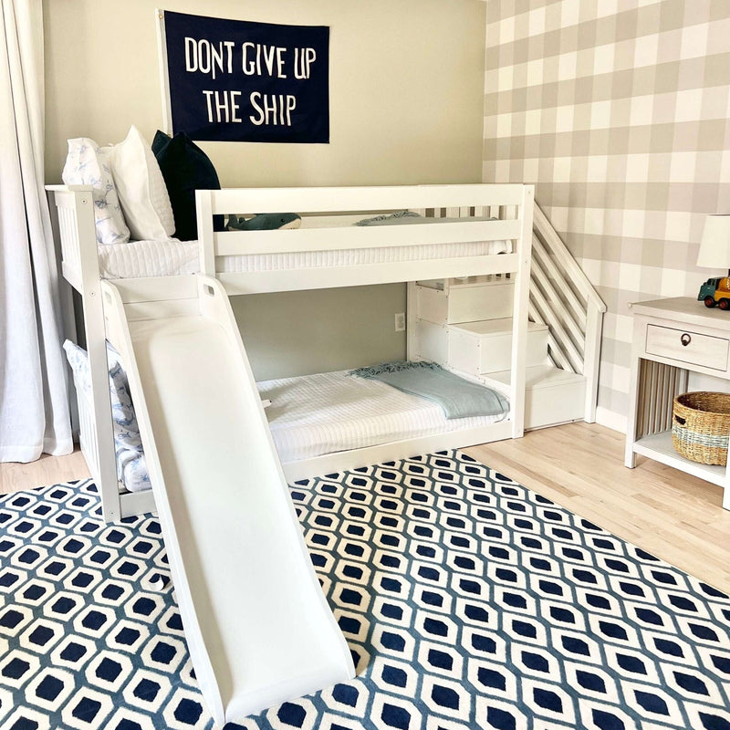 White kids bunk bed with slide in nautical nursery, navy "Don't Give Up The Ship" wall banner, blue geometric rug and neutral plaid accent wall