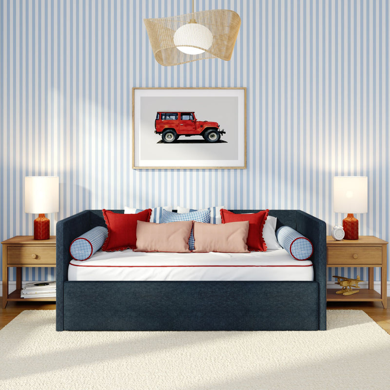 Navy upholstered daybed with red and pink throw pillows against blue-and-white striped wallpaper, framed red vintage SUV wall art, wooden nightstands and lamps