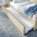 Pull-out trundle bed drawer beneath modern upholstered beige bed with white linens and blue throw blanket on patterned rug