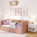 Cozy pink girl's bedroom with blush upholstered daybed, butterfly pillow, lavender bedding, floral framed prints, white nightstand with lamp and pink alarm clock