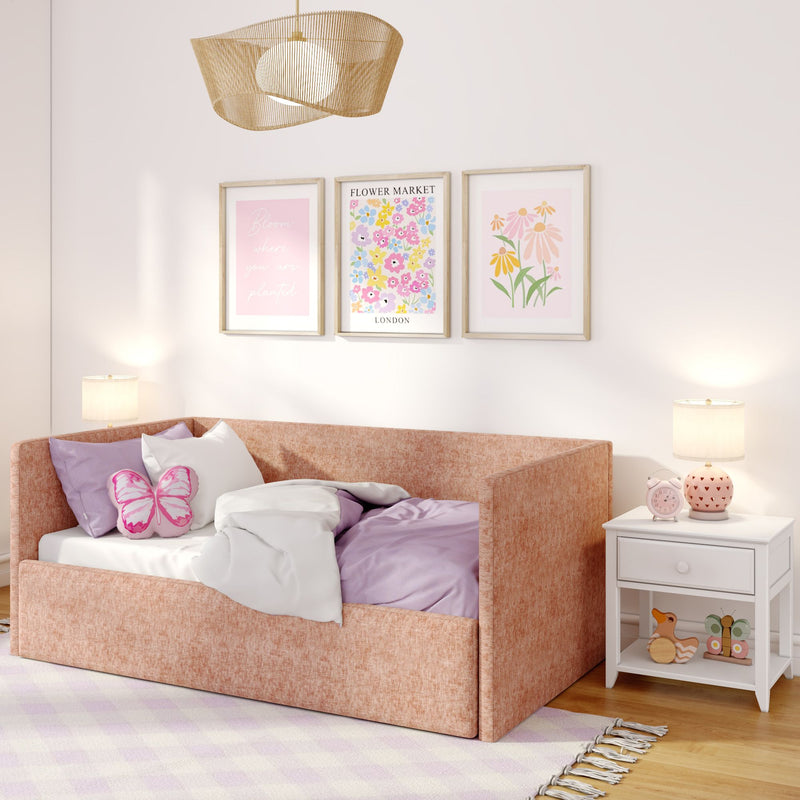 Cozy pink girl's bedroom with blush upholstered daybed, butterfly pillow, lavender bedding, floral framed prints, white nightstand with lamp and pink alarm clock